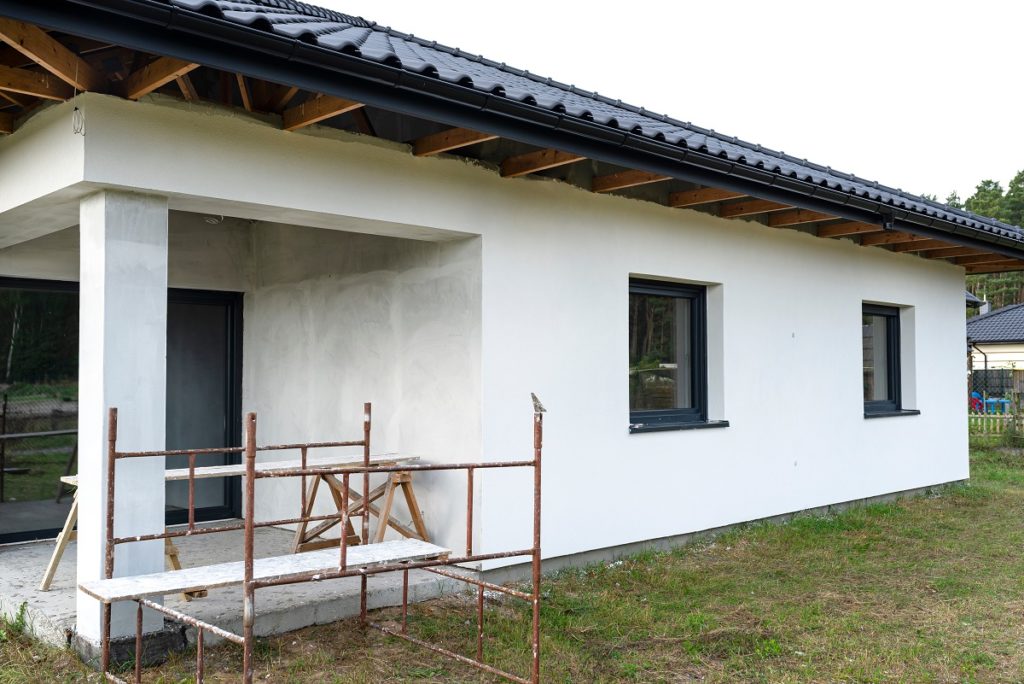 A newly applied layer of white silicone plaster on the wall of the house, visible metal scaffolding and boards.